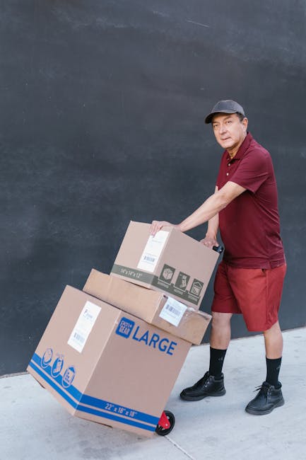 A delivery person dressed in a maroon shirt, red shorts, and black shoes is pushing a hand truck with two large cardboard boxes labeled 'LARGE' and bearing delivery barcodes, against a plain grey wall