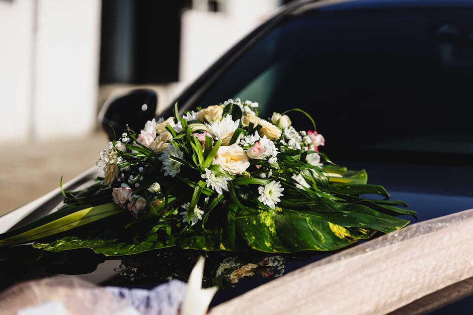 A floral arrangement featuring a bouquet of fresh white roses, daisies, and small pink blossoms, accented with lush green foliage. The bouquet is elegantly wrapped in clear cellophane with a subtle ri
