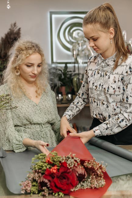 In the interior of a florist shop, two women are working together on a floral arrangement. The woman on the right, with light brown hair tied back and wearing a patterned blouse, is carefully wrapping