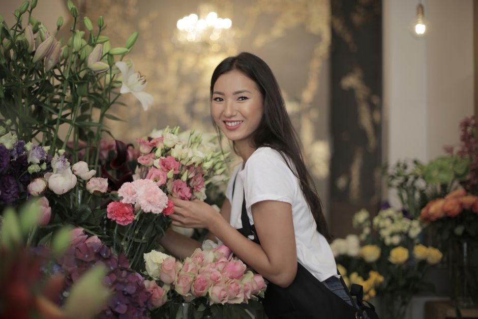 A young woman with long dark hair and a bright smile is selecting a floral gift in a florist shop, surrounded by an array of fresh flowers including pink roses, white lilies, and purple blooms, all ar