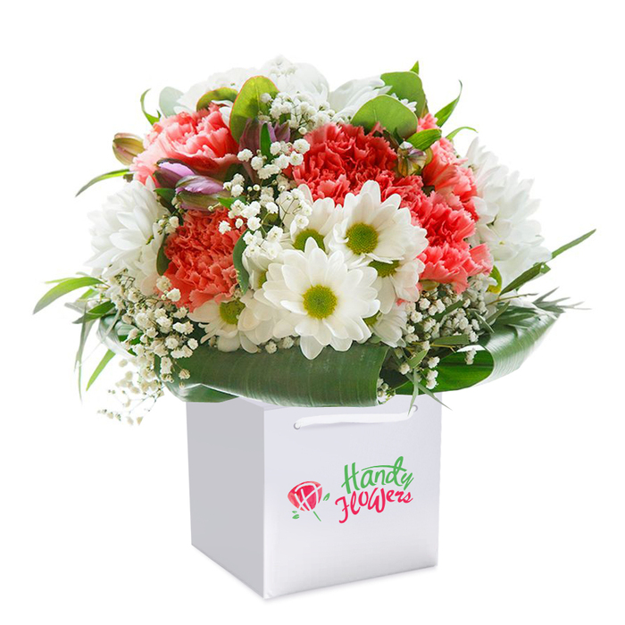 This radiant bouquet is a true celebration of floral artistry, expertly arranged by a seasoned Ickenham floral designer and displayed upright in a sleek, square white gift bag. The composition is anchored by a mix of lush coral-pink and deep red carnations, their ruffled, velvety petals layered for a rich, tactile effect. Dotted throughout are cheerful white daisies, each featuring a vivid yellow-green heart that draws the eye, and gentle sprays of ethereal white gypsophila, adding an airy lightness to the overall design. Facing outward, the foliage is abundant and deep green, wrapping the stems in a glossy embrace reminiscent of the shaded lanes in Ickenham's village heart. Delicate, unopened blooms tinged in purple or pink lend the arrangement depth and a sense of anticipation-a nod to new beginnings celebrated in this West London suburb. The spotless white bag is neatly finished with soft ribbon handles, an echo of attentive care and professionalism characteristic of local florists. Whether destined for a birthday gathering in a family home by Hillingdon Station or a thoughtful gift for a loved one recuperating at Hillingdon Hospital, this bouquet symbolises freshness, vibrant beauty, and the personal touch synonymous with flower delivery throughout Ickenham.