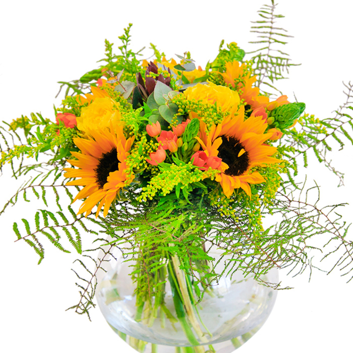 This exquisite bouquet, presented in a classic round glass vase, reflects the vibrant heart of Ickenham in every detail. Radiant sunflowers bloom at the center, their bold yellow-orange petals encircling espresso-hued centers, capturing the look of sunlight filtering through the trees in Ickenham's community gardens. Nestled alongside are lush yellow garden roses, each unfurling in tightly ruffled spirals, their velvety petals releasing the subtle scent of a summer evening after rainfall. Between these showstoppers, clusters of glossy red hypericum berries glimmer, providing lively flashes of colour much like the local berries found near Warren Road in peak season. Fronds of emerald fern flow gently around the bouquet, softening its silhouette and adding organic movement, as if shaped by a light Chiltern breeze. Dainty stalks of goldenrod and silver-green eucalyptus leaves peek through, blending into the mixed greenery with delicate charm. The clear glass vase-reminiscent of a fishbowl-provides a window into the arrangement's architectural grace, showcasing sturdy green stems neatly submerged in water. This bouquet is perfect for marking milestones, whether it's a birthday, anniversary, or the arrival of a loved one home in Ickenham. Every element is selected and arranged with the practiced touch of a local florist, blending artistry with warmth to create a display that radiates happiness and a true sense of place.