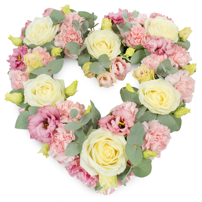 A luxurious, heart-shaped wreath for a funeral or memorial service is captured against a bright white backdrop, allowing its pastel tones to glow softly. The entire heart is made up of flowers packed closely together, forming a complete, symmetrical outline that feels steady and reassuring. Large cream and pale yellow roses are placed thoughtfully throughout the design, their spiralled centres and velvety outer petals giving a sense of quiet elegance and dignity. Around them, generous clusters of soft pink carnations add volume and texture; their many, slightly crimped petals overlap like delicate ruffles, creating a gentle, cloud-like surface. Interwoven with these are deeper pink to mauve lisianthus blooms, some more open and revealing darker centres, others still partly cupped, giving subtle movement and depth to the composition. Fine sprays of yellow-green buds peek out between the larger flowers, hinting at freshness and new growth, while strands of silvery-green eucalyptus thread through the heart. The eucalyptus leaves, rounded and slightly frosted in tone, provide a cool contrast to the warm pastels and help define the shape, much as quiet tree-lined streets frame the character of Ickenham's village centre. The flowers are arranged in a way that feels both precise and tender, reflecting the hand of an experienced florist attentive to every stem. The overall impression is one of serene remembrance, ideal for resting beside a coffin, adorning a chapel of rest near Ickenham, or marking a place of reflection at a graveside, where family and friends gather to honour cherished memories and say a gentle farewell.